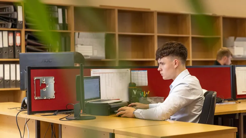 Mladý muž v bílé košili pracuje u stolního počítače v kanceláři s regály na šanony