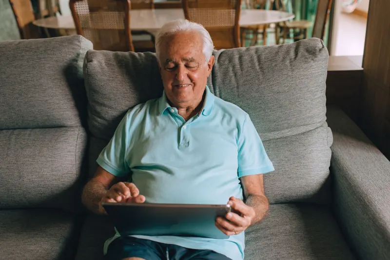 Starší muž v obývacím pokoji s úsměvem ovládá tablet na klíně